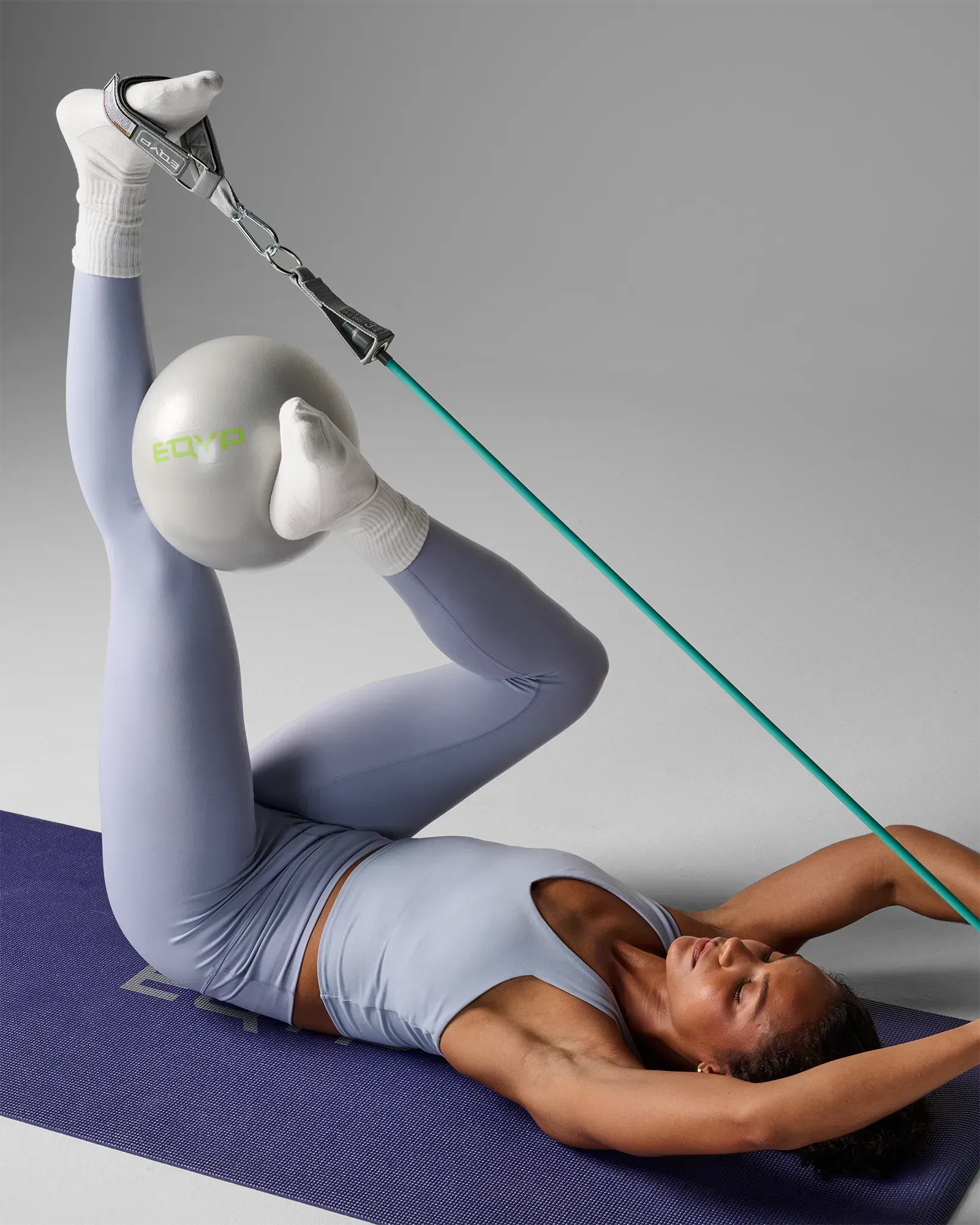 Woman performing a supine single-leg raise with ankle strap attachment using EQYP Power Tubes 40 lbs teal resistance tube on a yoga mat, with EQYP pilates ball between knees
