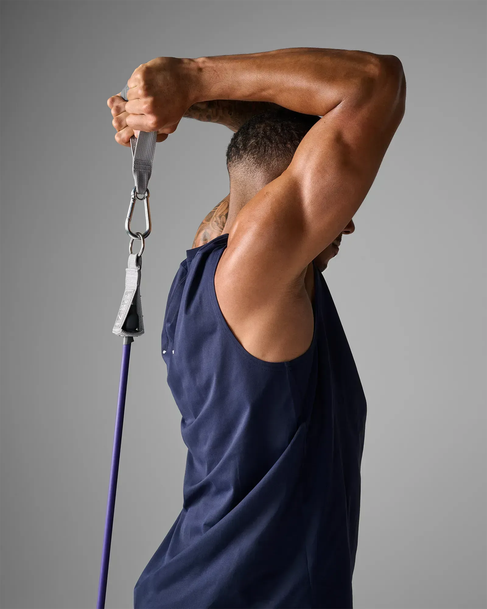 Man performing a standing unilateral overhead tricep extension using EQYP Power Tube 80 lbs purple resistance tube with gray handle, rear view