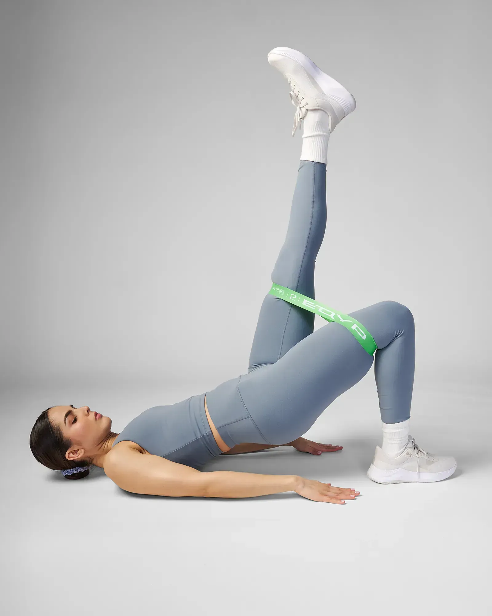 Woman performing a supine unilateral glute bridge with leg extension using EQYP Power Loop Medium green resistance band above the knees