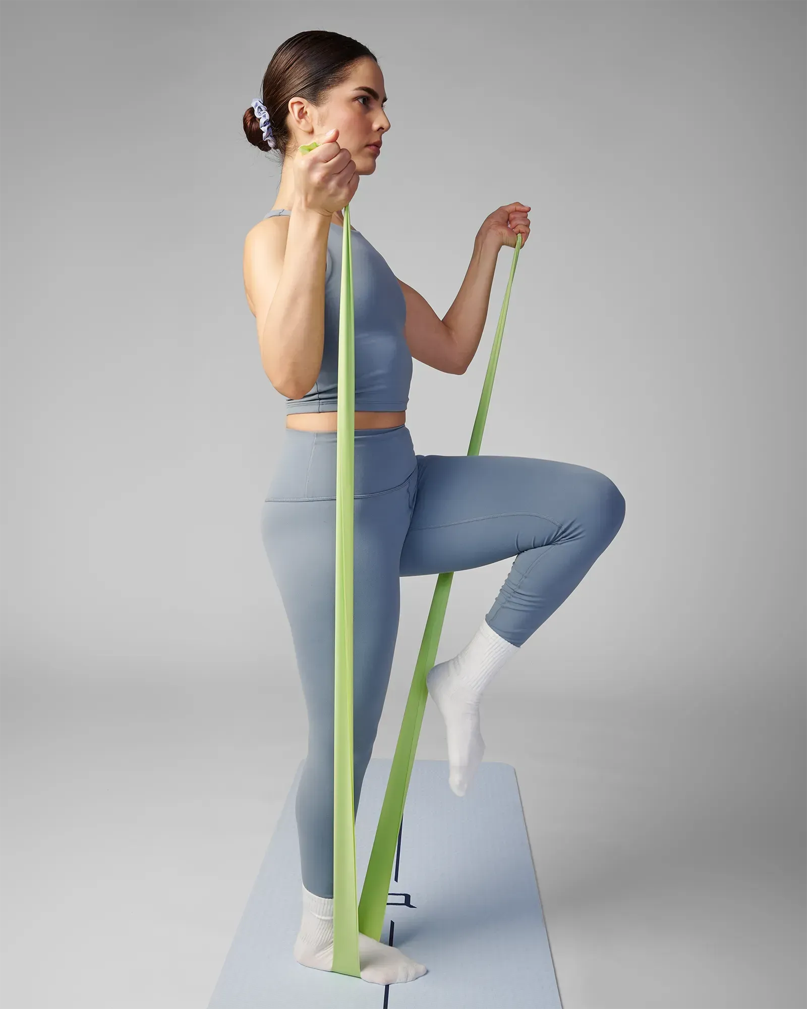 Woman performing a standing bicep curl with a knee raise on a yoga mat using an EQYP green latex-free flat resistance band