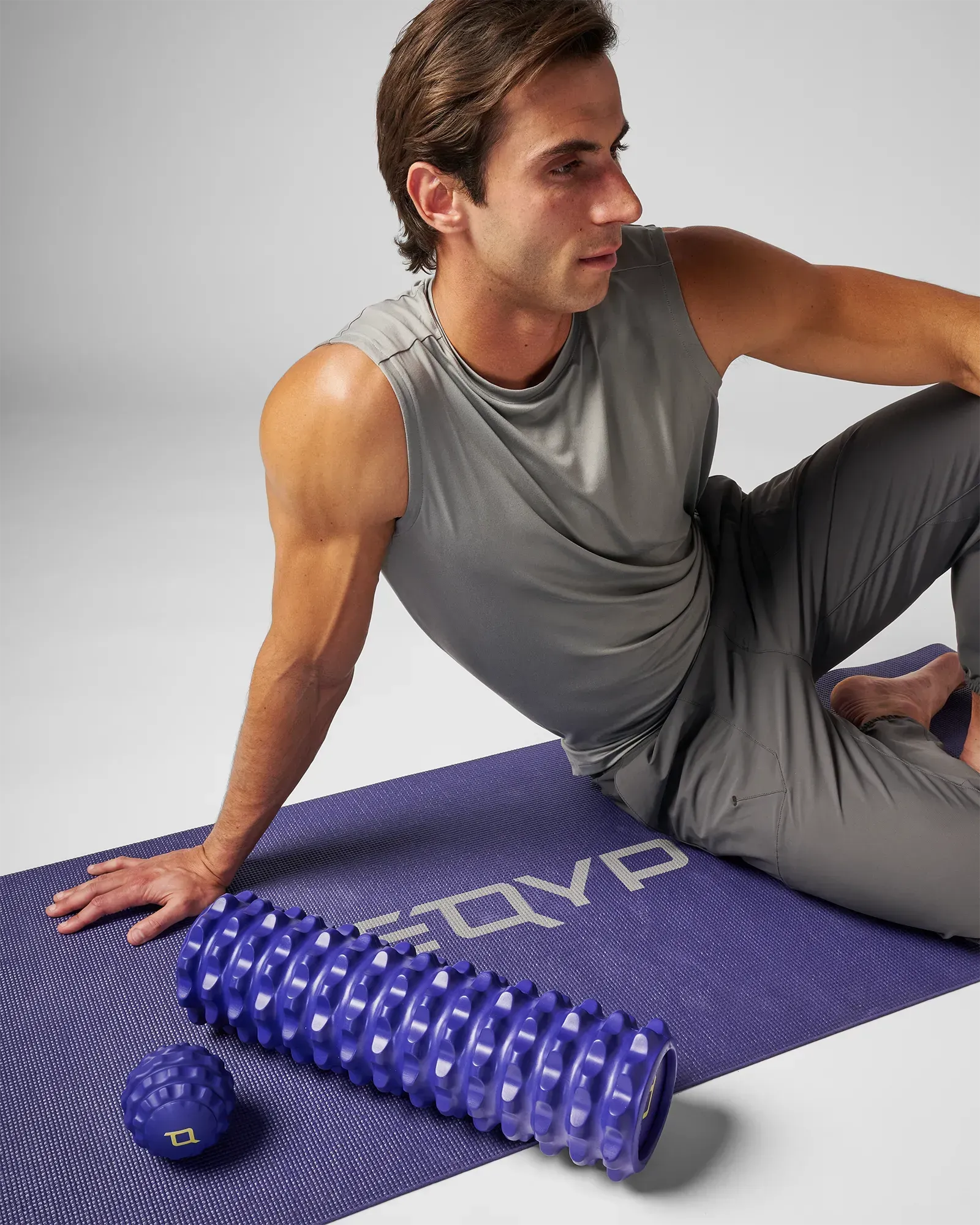 Man sitting on yoga mat next to medium sized blue EQYP massage ball and extreme 18 Inch EQYP Foam Roller