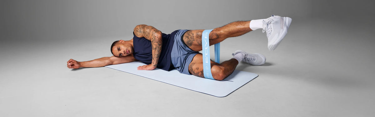 Man lying on a mat with his leg raised, using a light blue EQYP resistance band around his thighs for a full body strength exercise.
