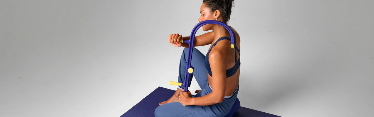 Woman using a purple and yellow EQYP muscle hook tool to massage her upper back and shoulder while seated.