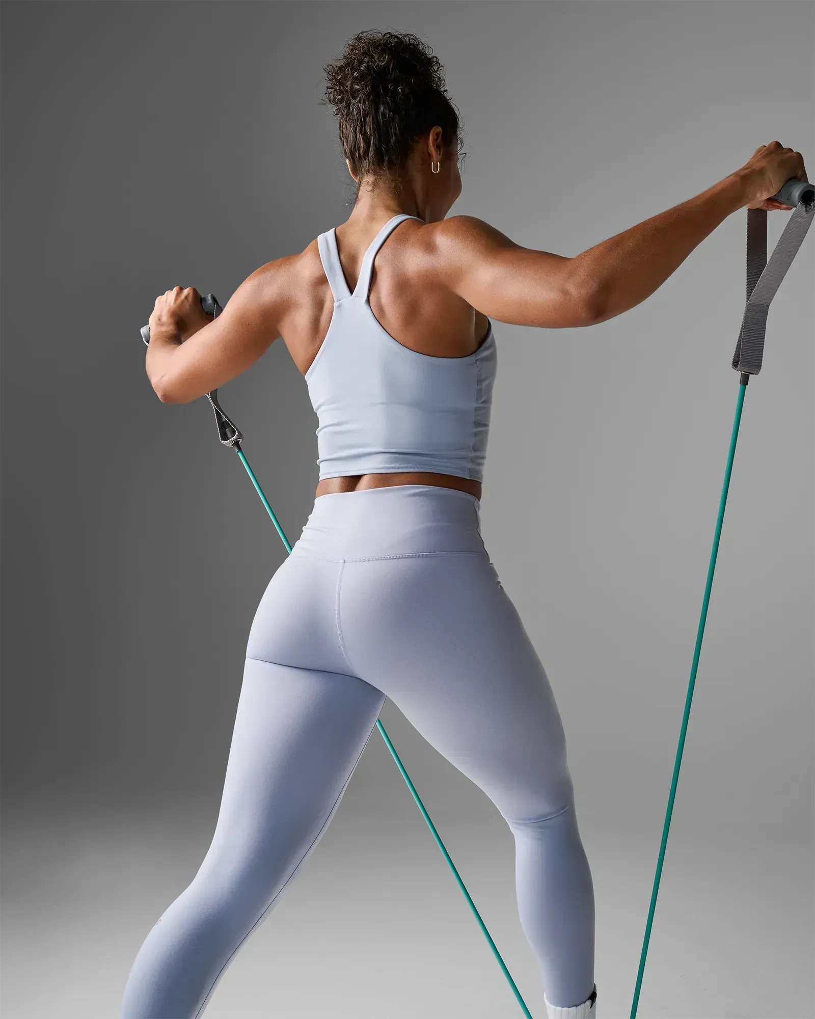 Woman performing a standing bilateral lateral raise using EQYP Power Tubes with Handles Medium teal resistance tube, rear view