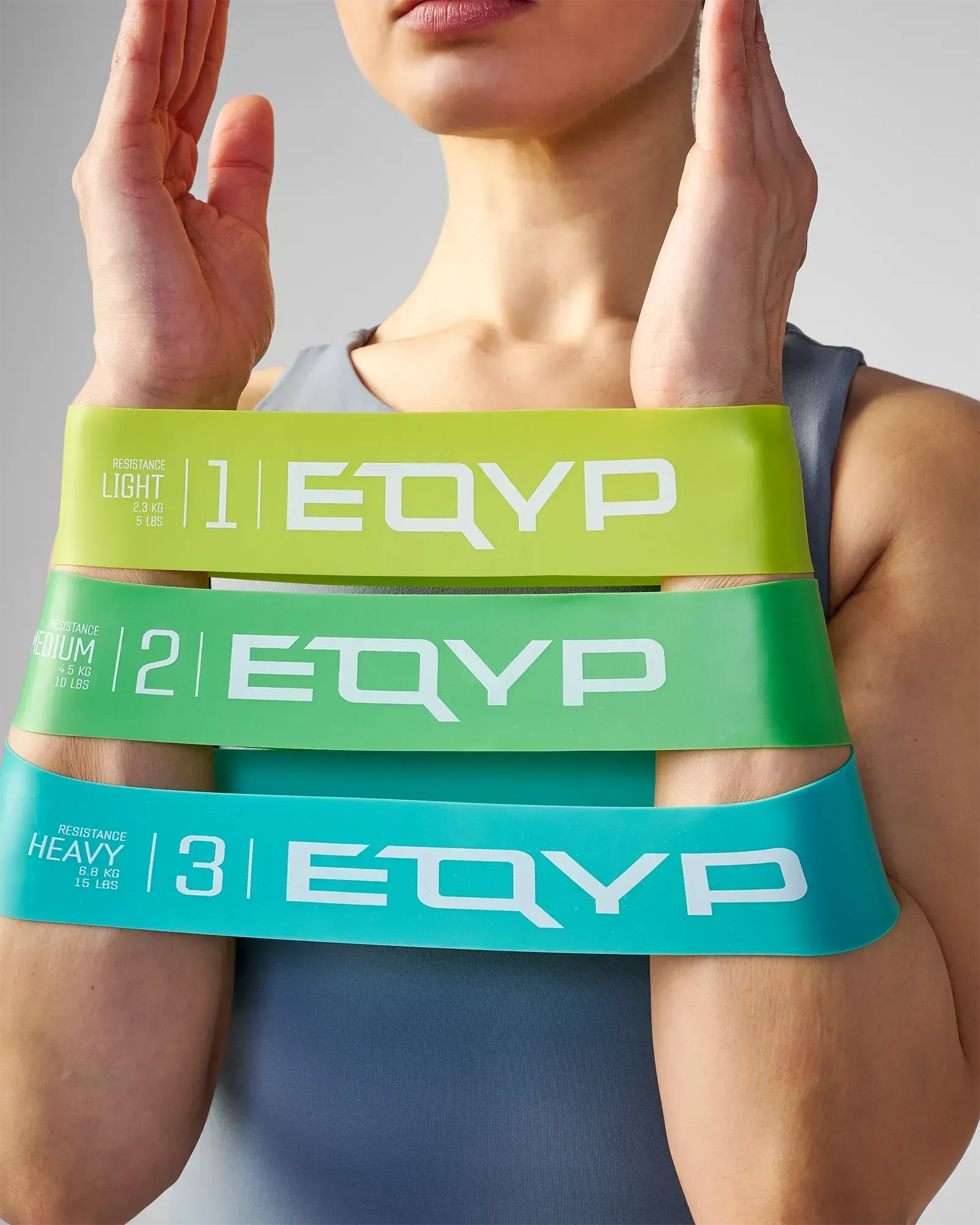 Woman holding all three EQYP Power Loops showing Light 5 lbs yellow-green, Medium 10 lbs green, and Heavy 15 lbs teal resistance bands